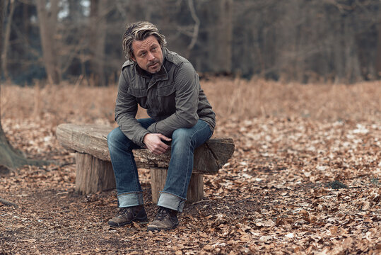 Blonde Man In Green Coat Sits On A Wooden Bench In Nature Reserve.