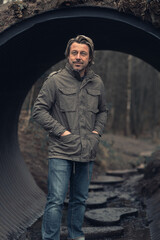 Blond man in green jacket and jeans stands in a tube tunnel in nature reserve.