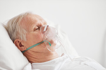 Side view portrait of ill senior man lying in hospital bed with oxygen supplementation mask , copy...