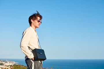Fototapeta premium A Caucasian man from Spain standing on a grassy hill next to the ocean with a black purse over his shoulder