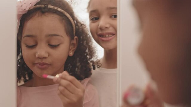Chest-up of reflection of two little Mixed-Race sisters looking in mirror, experimenting with makeup at home. Younger kid applying lip gloss, smiling, older girl kissing and talking to her