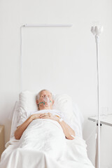 Vertical portrait of ill senior man lying in hospital bed with oxygen supplementation mask and eyes...