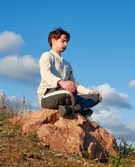 Naklejka na ściany i meble A vertical shot of a Caucasian man from Spain sitting on a rock surrounded by grass and meditating on sky background