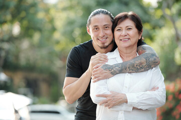 Portrait of handsome mixed-race adult man hugging his mature mother from behind and smiling at camera when they are standing outdoors