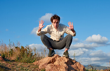 A Caucasian man from Spain putting his hands up while squatting on a rock surrounded by grass
