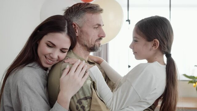 A soldier man is holding on his hands his young daughter while his wife is hugging his back at home
