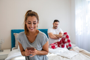 Addicted young woman on bed texting while man looks angry. Young couple having a problem. Woman is chatting using a smartphone while her boyfriend is bored in bed