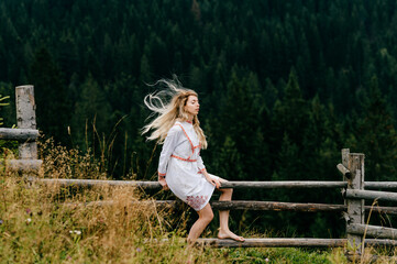 Naklejka na ściany i meble Young attractive blonde girl in white dress with embroidery sitting on wooden fence over picturesque forest landscape