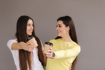 Two attractive girls hold cup of coffee in studio on grey background. Brunette female in yellow sweater. Beautiful females smile