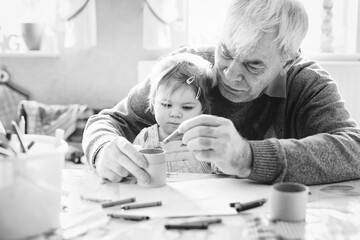 Cute little baby toddler girl and handsome senior grandfather painting with pencils at home. Grandchild and man having fun together. Family and generation in love. Old black and white image