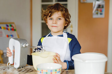Adorable funny blond little kid boy baking chocolate cake and tasting dough in domestic kitchen, indoors. Happy child having fun with working with mixer, flour, eggs dough at home. Little helper