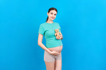 Happy pregnant woman holding a teddy bear against her belly at Colored background. Young mother is expecting a baby. Copy space