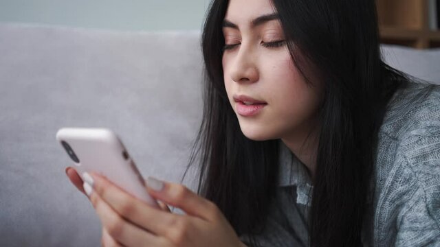 A thinking young asian woman is using her smartphone while lying on the sofa at the apartments