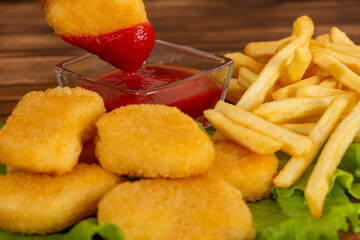 Juicy chicken nuggets are dipped in ketchup. Nuggets and French fries on a wooden background.