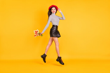 Full size profile side photo of lady jump up walk hold tulips wear leather skirt striped turtleneck isolated on yellow color background