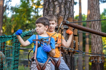 Two cute happy young children, boy and his brother in protective harness, carbine and safety helmets on rope way on bright sunny day on green foliage bokeh background.