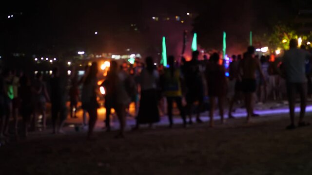 Crowded beach at night time, people come for to dance party and watch performance, blurred view. Spectators silhouettes on foreground, woman dance, fire twirler with pois, long jump rope on background