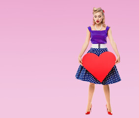 Full body of woman holding heart symbol, dressed in pin up style dress with polka dot, over pink...