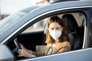 Young woman driving car with protective mask on her face. Looking worried at camera. Healthcare, virus protection, allergy protection concept. 