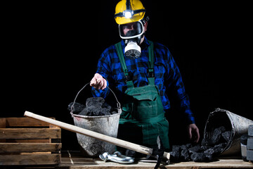 A miner wearing a helmet with a flashlight and a dust mask holds a bucket full of black coal in his hands