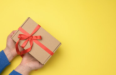 female hands hold brown cardboard box with red ribbon on yellow background, top view