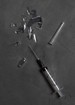 Used disposable syringe and broken glass ampoule with a dose of medication on a gray concrete background.