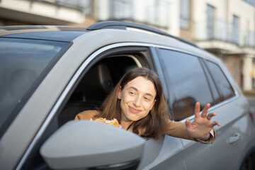 Happy woman eating a burger in the car while greet someone on the road. Have unhealthy fast food snack. Food to go. Hungry and busy concept.