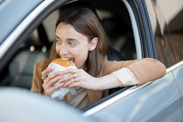 Happy woman eating a burger in the car. Bites a sandwich. Have unhealthy fast food snack. Food to...