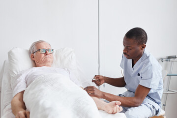 Portrait of African-American male nurse caring for senior man lying in hospital bed againts white, copy space