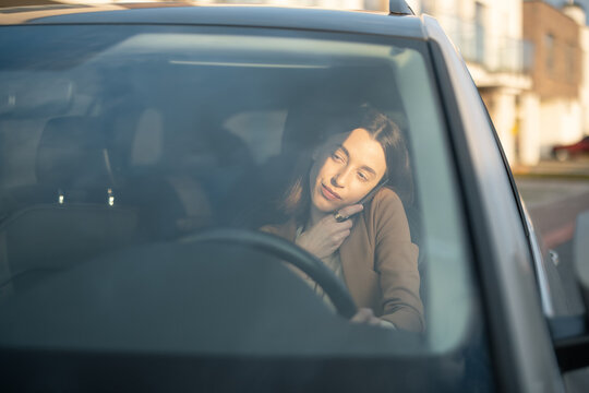 Young Woman Talking On The Phone While Driving A Car And Holding A Phone On Her Shoulder. Businesswoman Going To Work From The House. View Through The Windshield.