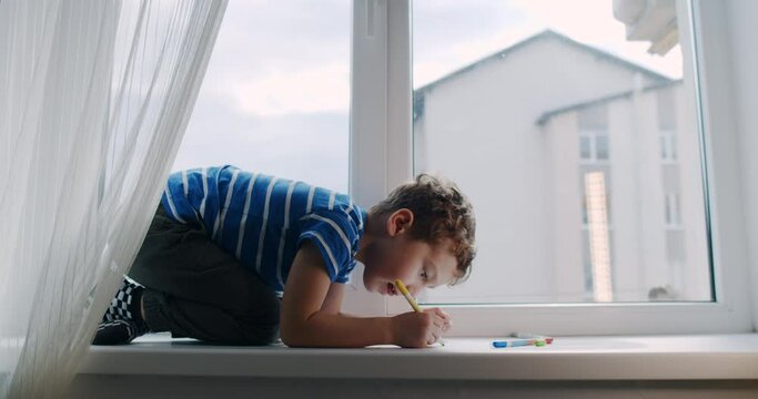 A small child, a happy boy, learns to draw on the window sill at home.