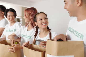 Pretty young smiling woman volunteering with her friends at donation center and packing groceries...