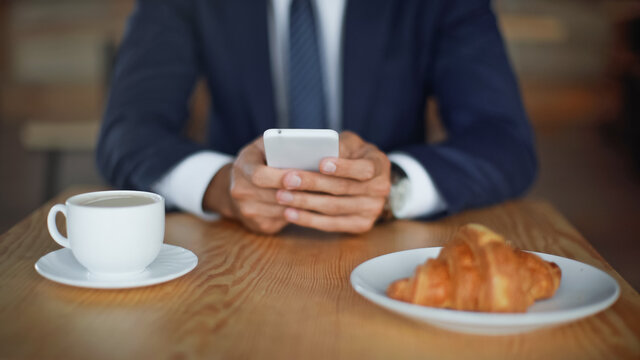 cropped view of businessman using smartphone near croissant and coffee in cup on blurred foreground