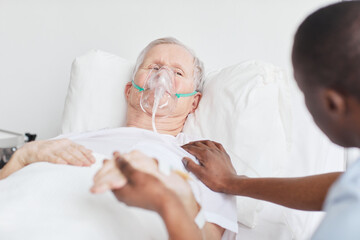 Portrait of senior man lying in hospital bed with oxygen mask and holding hands with caring doctor...