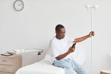 Portrait of young African-American man using smartphone in hospital room with iv drip setup, copy space