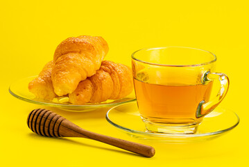 Freshly prepare croissant in a glass plate a cup of tea and a wooden honey dipper on yellow background.