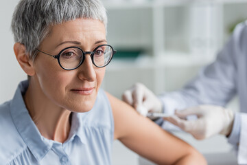 Mature woman sitting near doctor during vaccination on blurred background