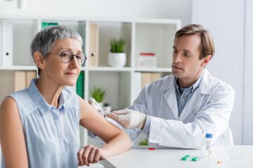 Doctor doing vaccination of mature woman near pills and vaccine on blurred foreground