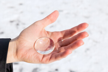 Small round transparent glass lens in the hand of a man against a background of snow. Find concept. Winter