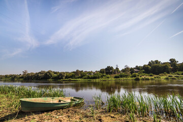 Obraz premium Lonely old boat on the river bank. Tall green grass grows. In the background there is a blue sky and low trees.