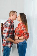 Loving man hugging his pregnant wife from behind standing near window at home, copy space