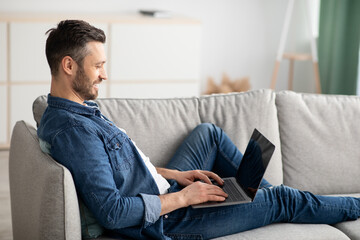 Side view of bearded man reclining on couch, using laptop