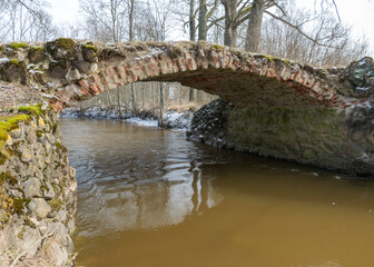 Fototapeta na wymiar a continuous boulder stone bridge with a brick used for masonry, early spring, bare trees, snow plan on the ground, Stone arch bridge over the river Kuja, Madona, Latvia