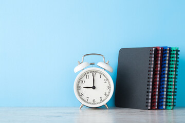 White alarm clock and notebooks on wooden table at light blue wall background. Planning time concept. Closeup. Empty place for text. Front view.