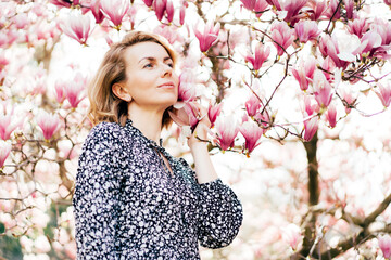 Young attractive confident woman in a dress in the park near a blooming magnolia tree inhales the aroma of flowers and enjoys the spring.