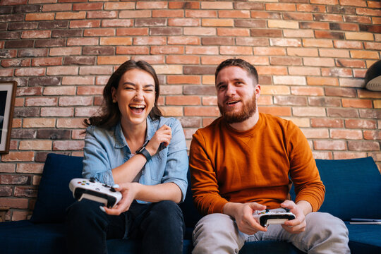 Front View Of Happy Laughing Young Couple Holding Controllers And Playing Video Games On Console Sitting Together On Couch At Cozy Living Room. Concept Of Leisure Activity Of Lovers At Home.