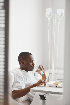 Vertical side view portrait of African-American man eating dinner in hospital while lying on white bed