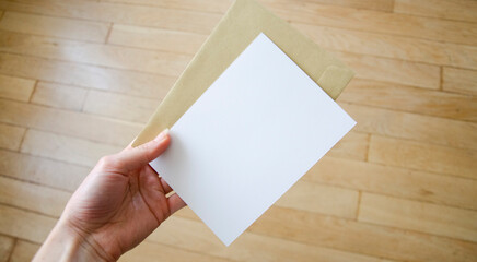 Hand holding greeting card with envelope, postcard, invitation, blank paper mock up on wooden background.