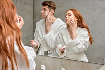 adorable caucasian couple take care of facial skin, woman applying cream while man is going to shave stubble