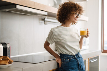 Smiling curly woman drinking juice while standing at home kitchen